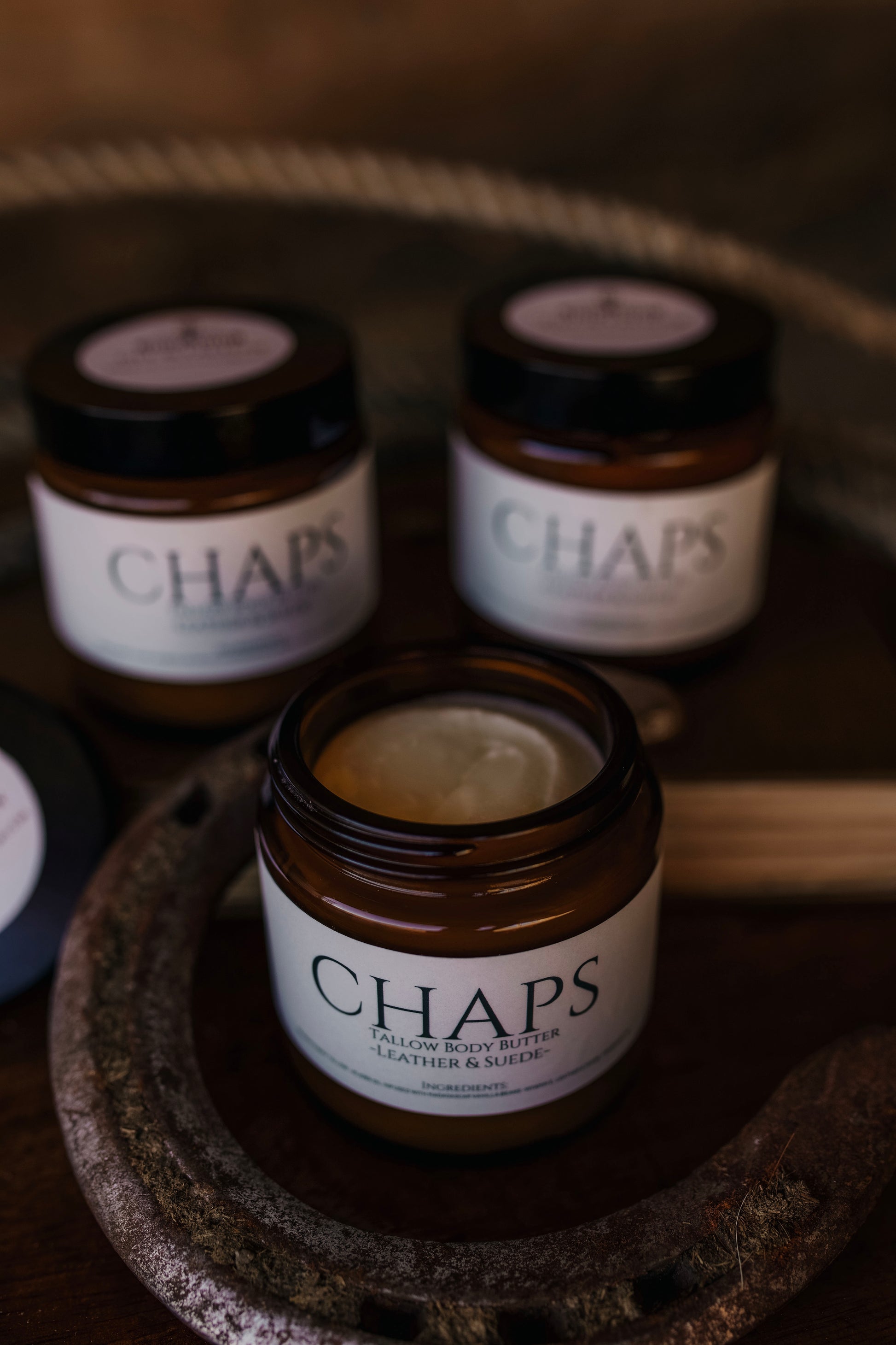Three jars of Till & Tallow CHAPS Leather & Suede body butter arranged on a textured wooden surface, with one open jar showing the body butter.