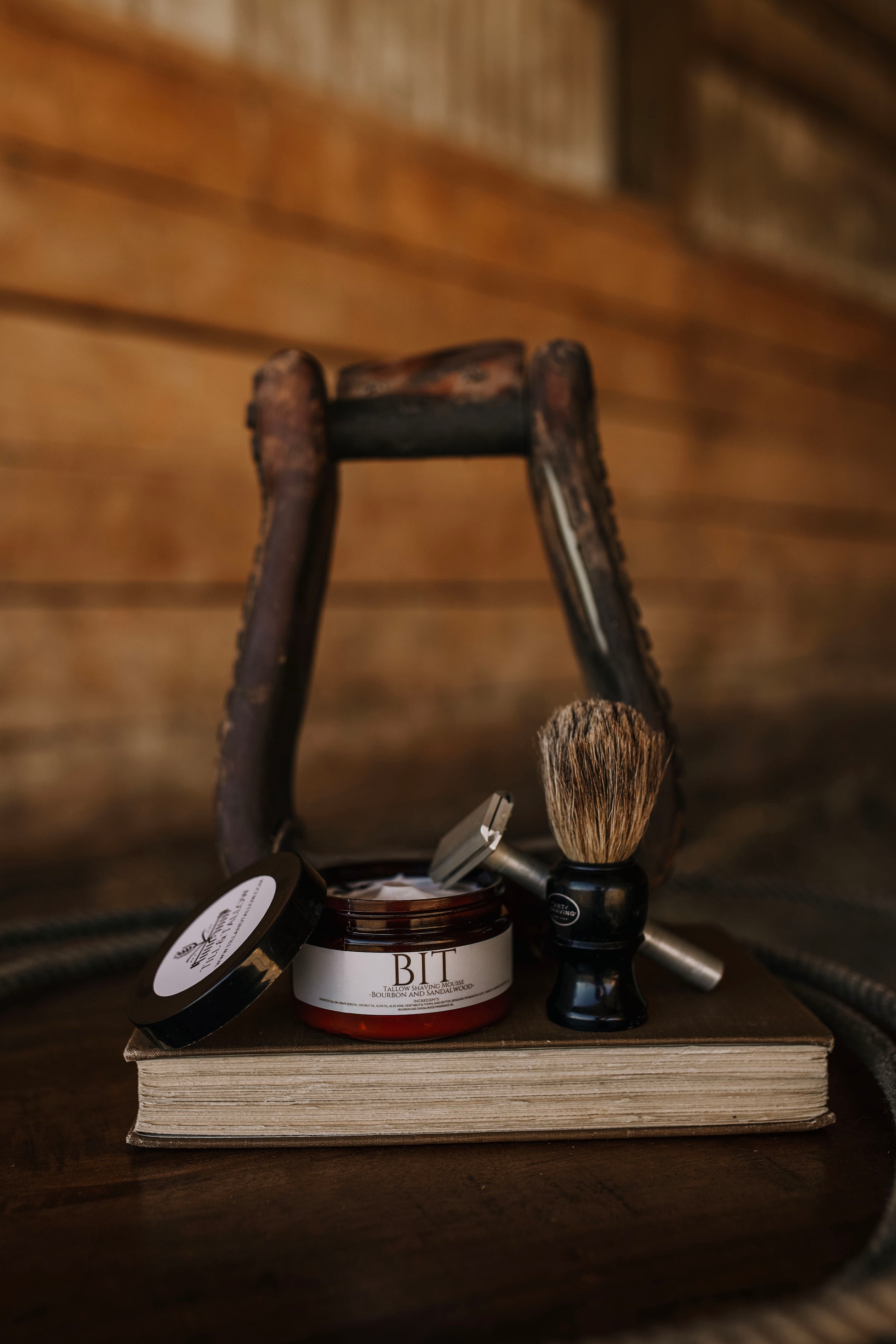 Till & Tallow Bit Shaving Mousse displayed with a shaving brush and razor inside a rustic horse stirrup, handcrafted in Alberta