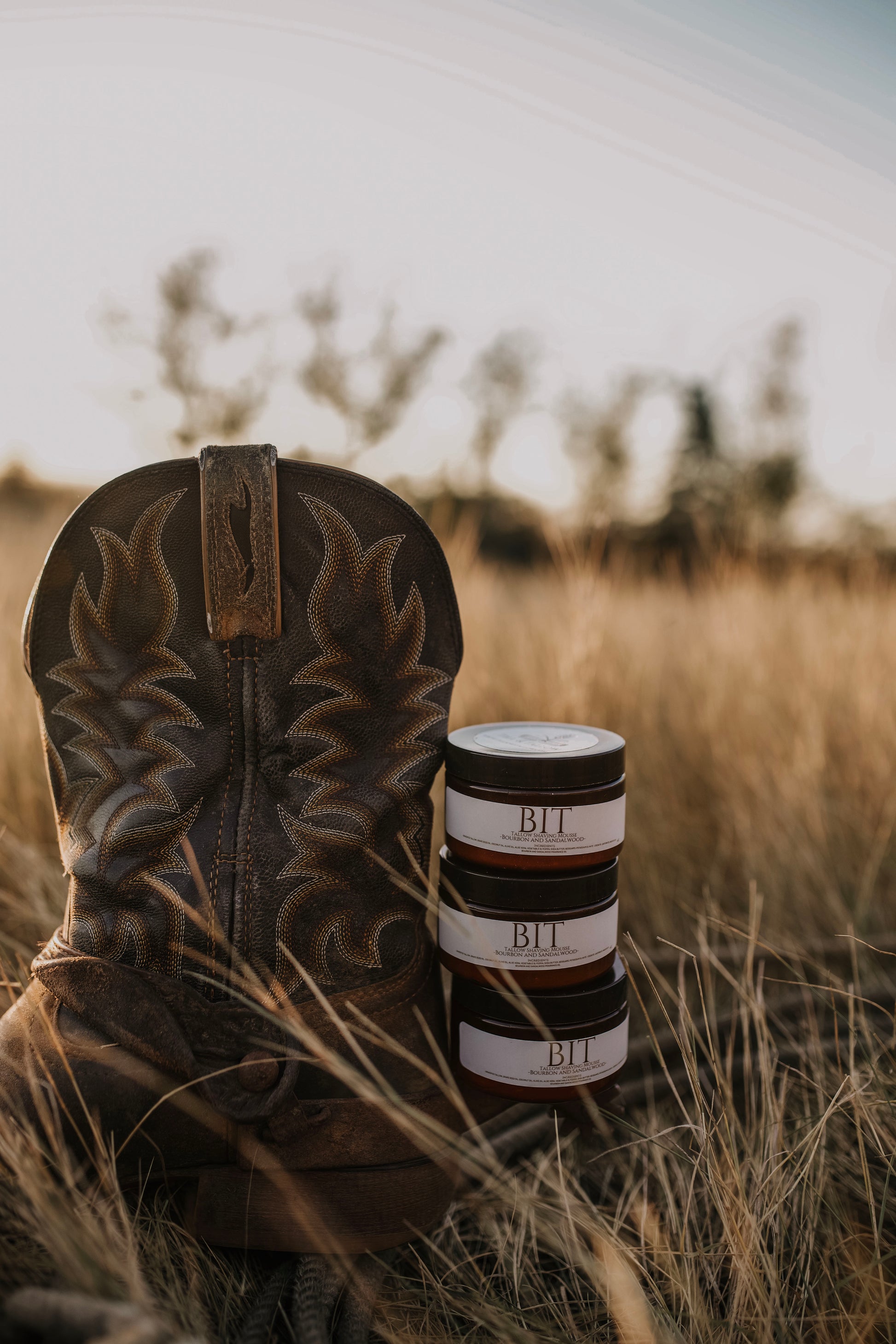 Till & Tallow Bit shaving Mousse jars stacked beside a brown cowboy boot in a grassy Alberta field, part of the Cowboy Line collection.