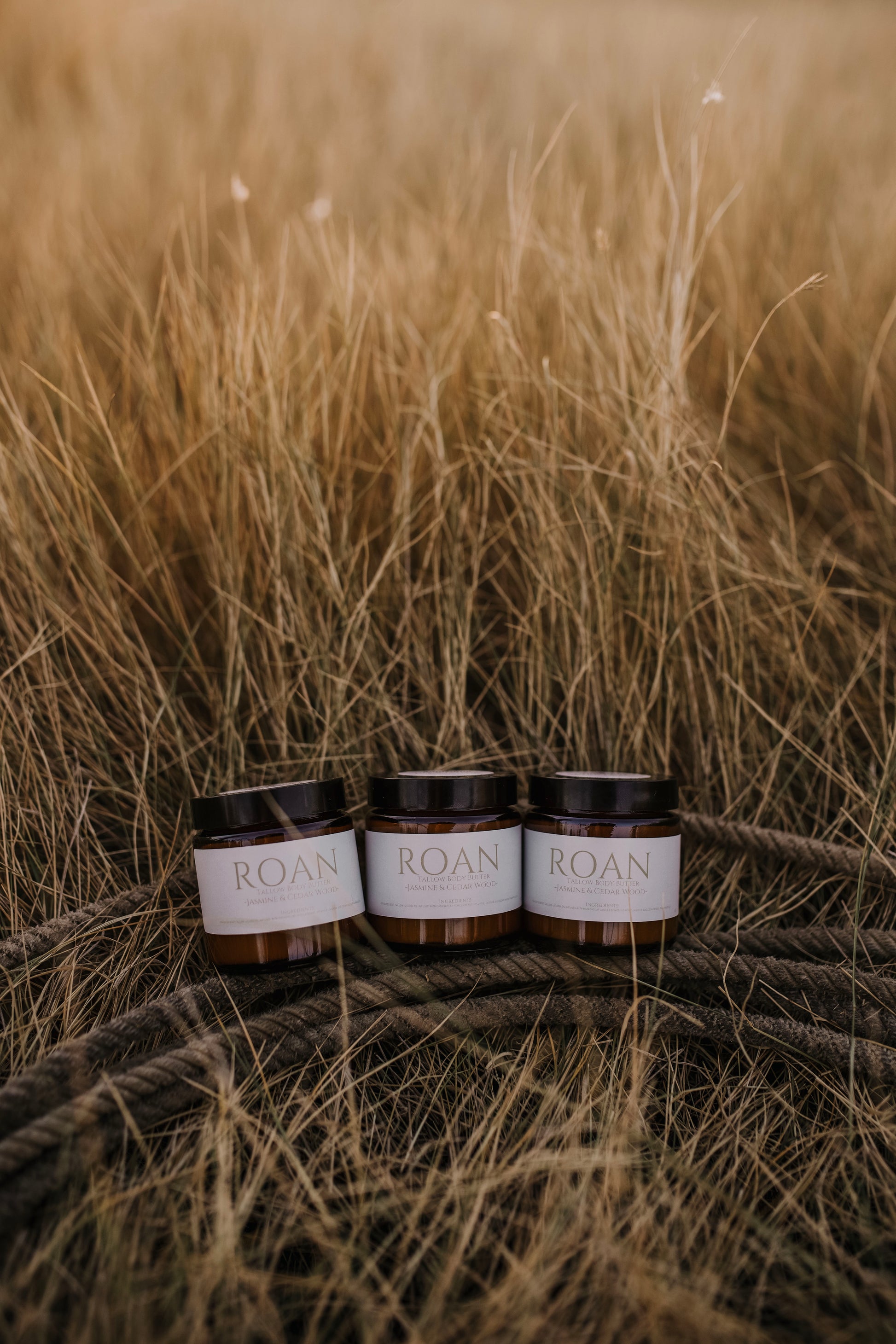 Till & Tallow Roan Body Butter jars beside a coiled cowboy rope in Alberta grass, photographed outdoors in natural light.