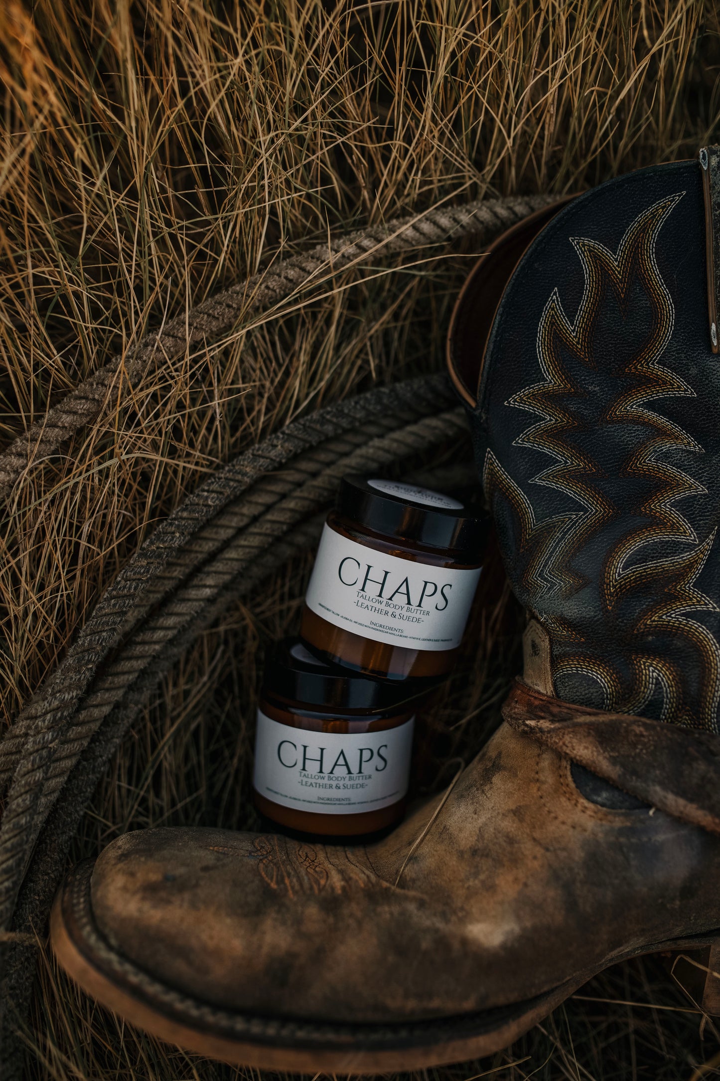 Till & Tallow Chaps Body Butter jars beside a worn cowboy boot in dry Alberta grass, rustic outdoor setting