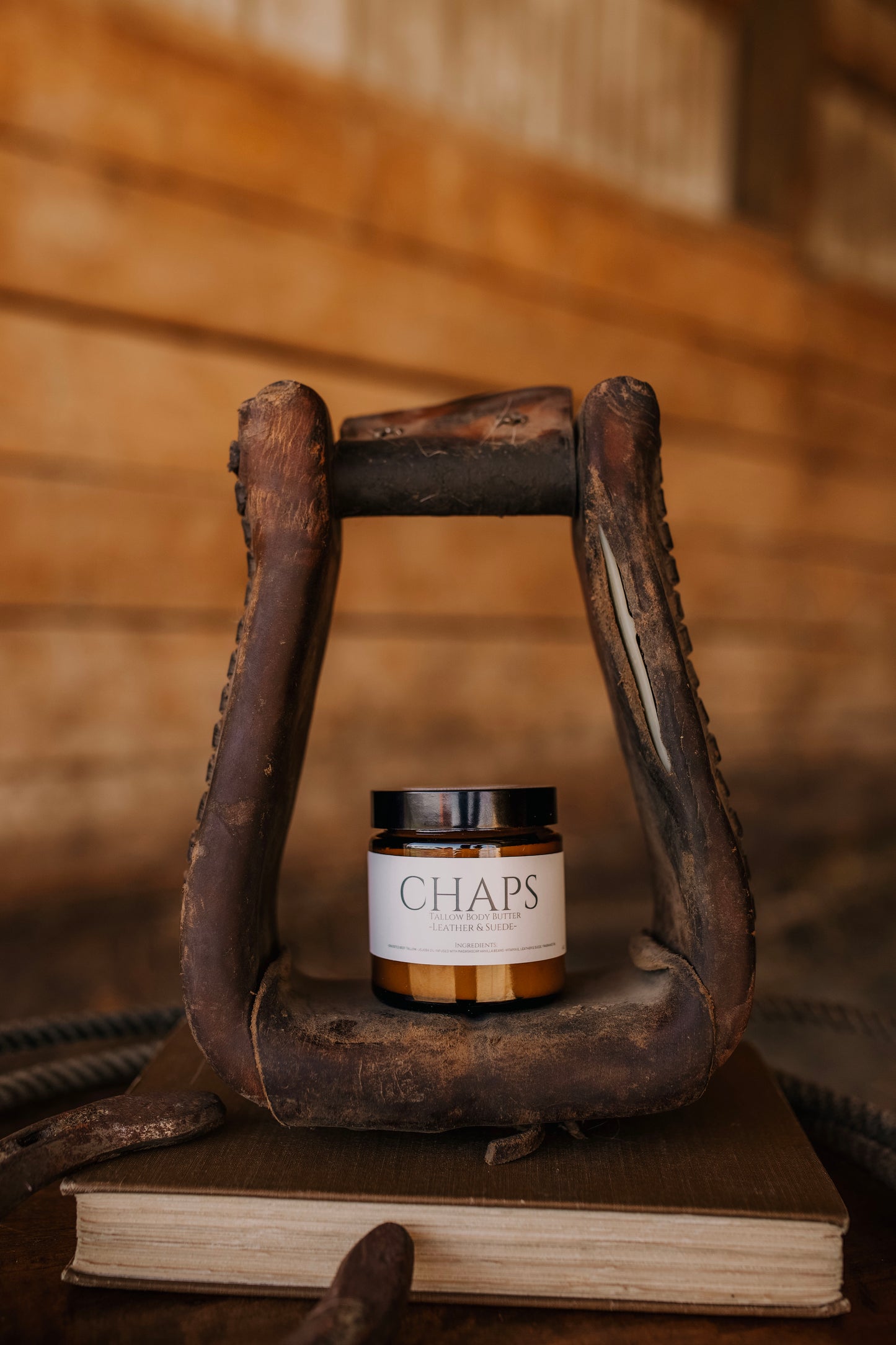 Till & Tallow Chaps Body Butter in amber jar displayed inside a rustic horse stirrup on a wooden surface, handcrafted in Alberta