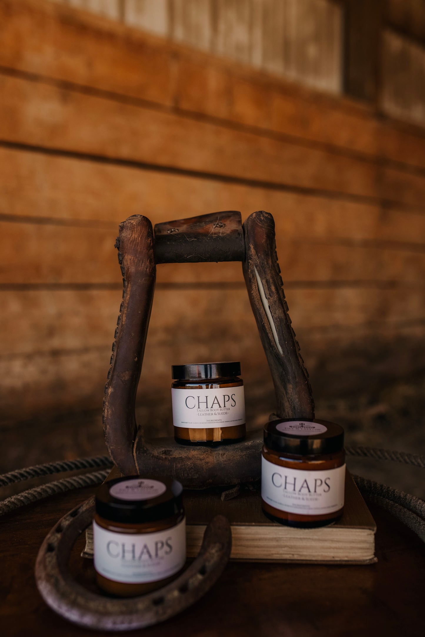 Till and Tallow chaps Body Butter jars arranged on a rustic wooden surface with a horse stirrup and warm Alberta Barn background.