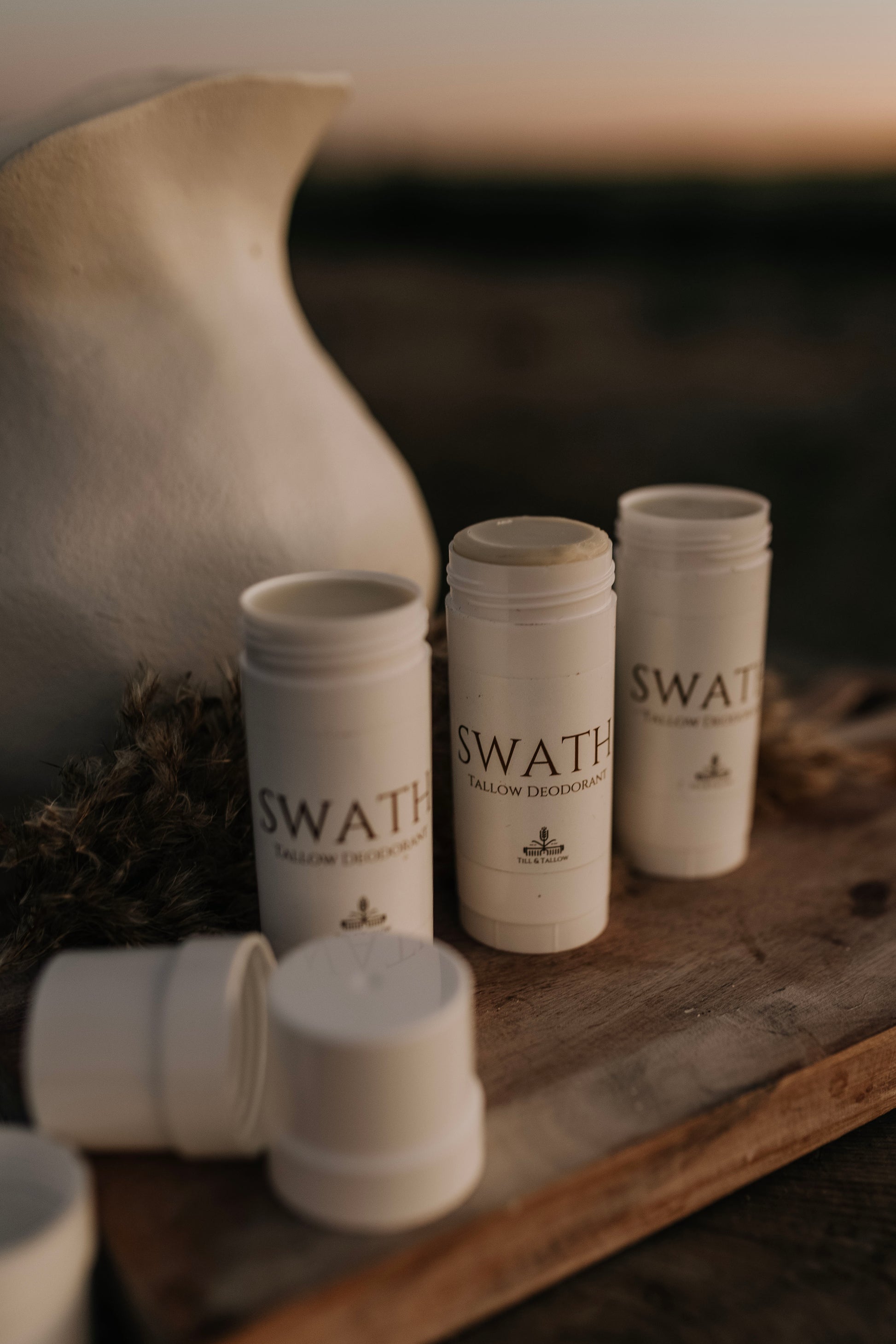 Till & Tallow SWATH natural deodorant tubes in white packaging displayed on a rustic wooden surface beside a white pitcher, handcrafted in Alberta.