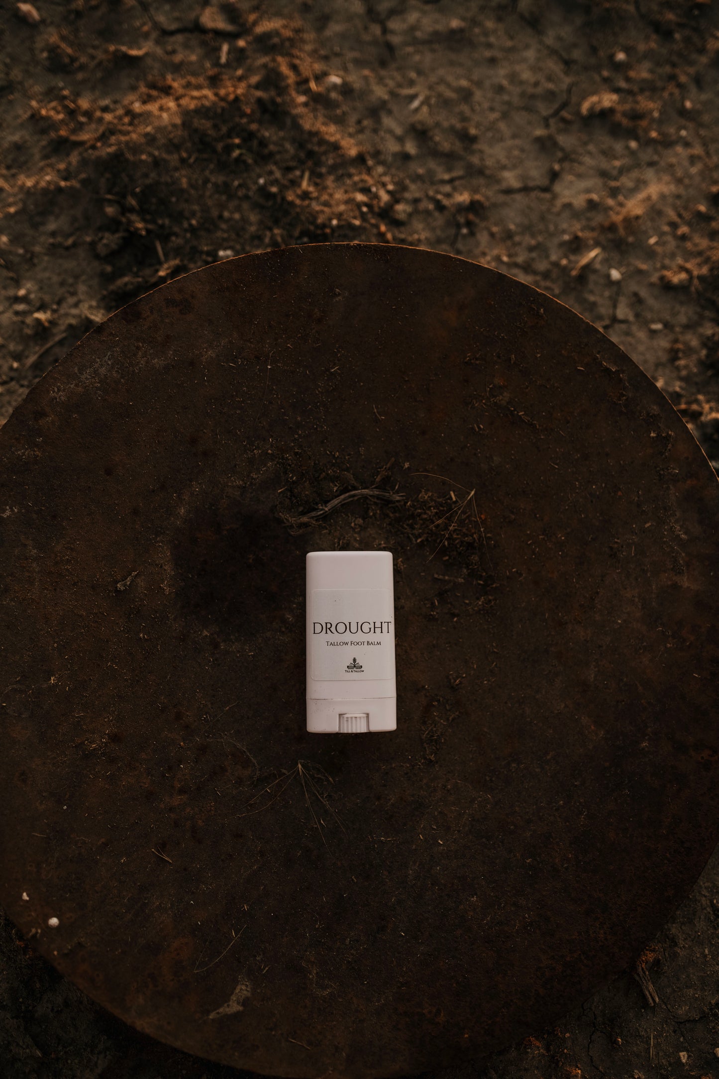 Till & Tallow Drought Cracked Heel Balm stick in white tube displayed on a rustic dark metal disk, handcrafted in Alberta, Canada