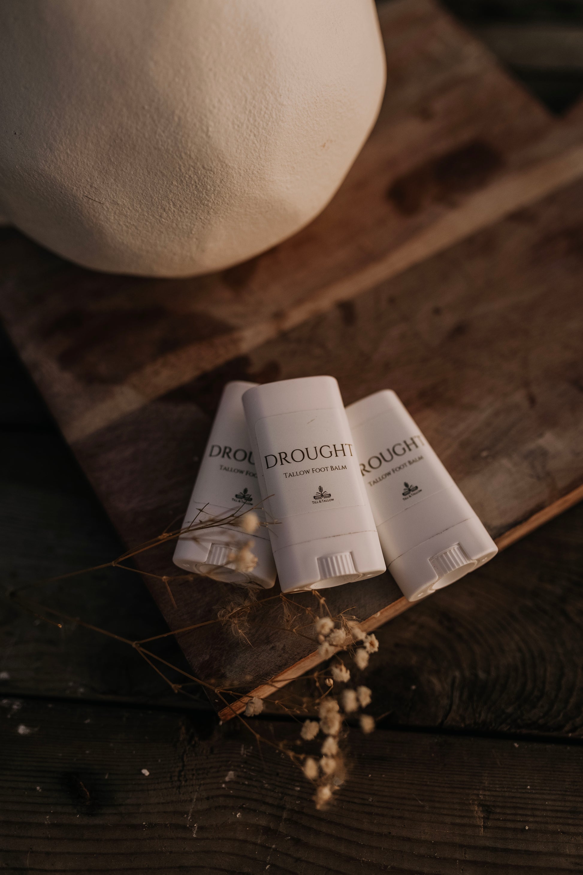 Three Till & Tallow Drought Cracked Heel Balm tubes arranged on a rustic wood with florals, handcrafted in Alberta
