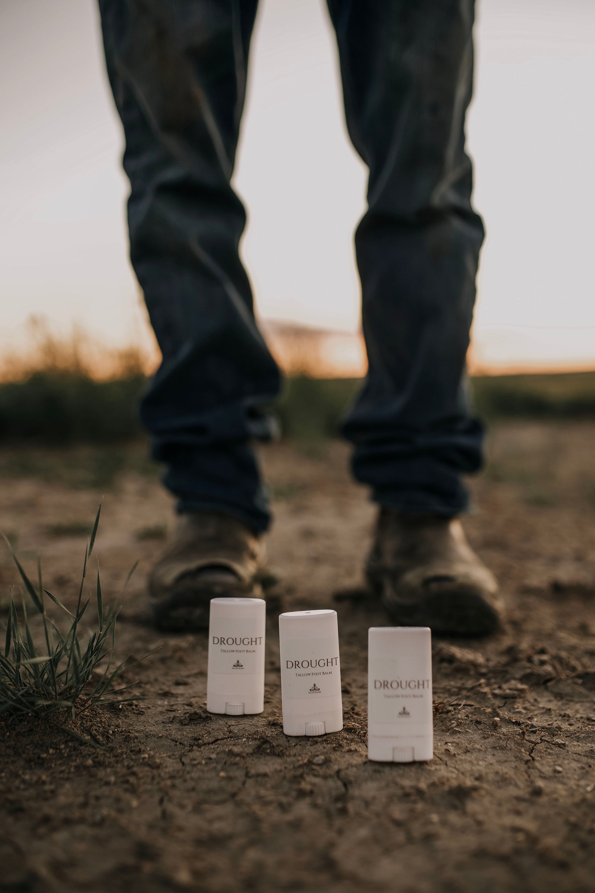 Till & Tallow Trio of Drought Cracked Heel Balm Stick tubes standing on earthy ground outdoors with a  farmer in jeans and boots in the background, handcrafted in Alberta.