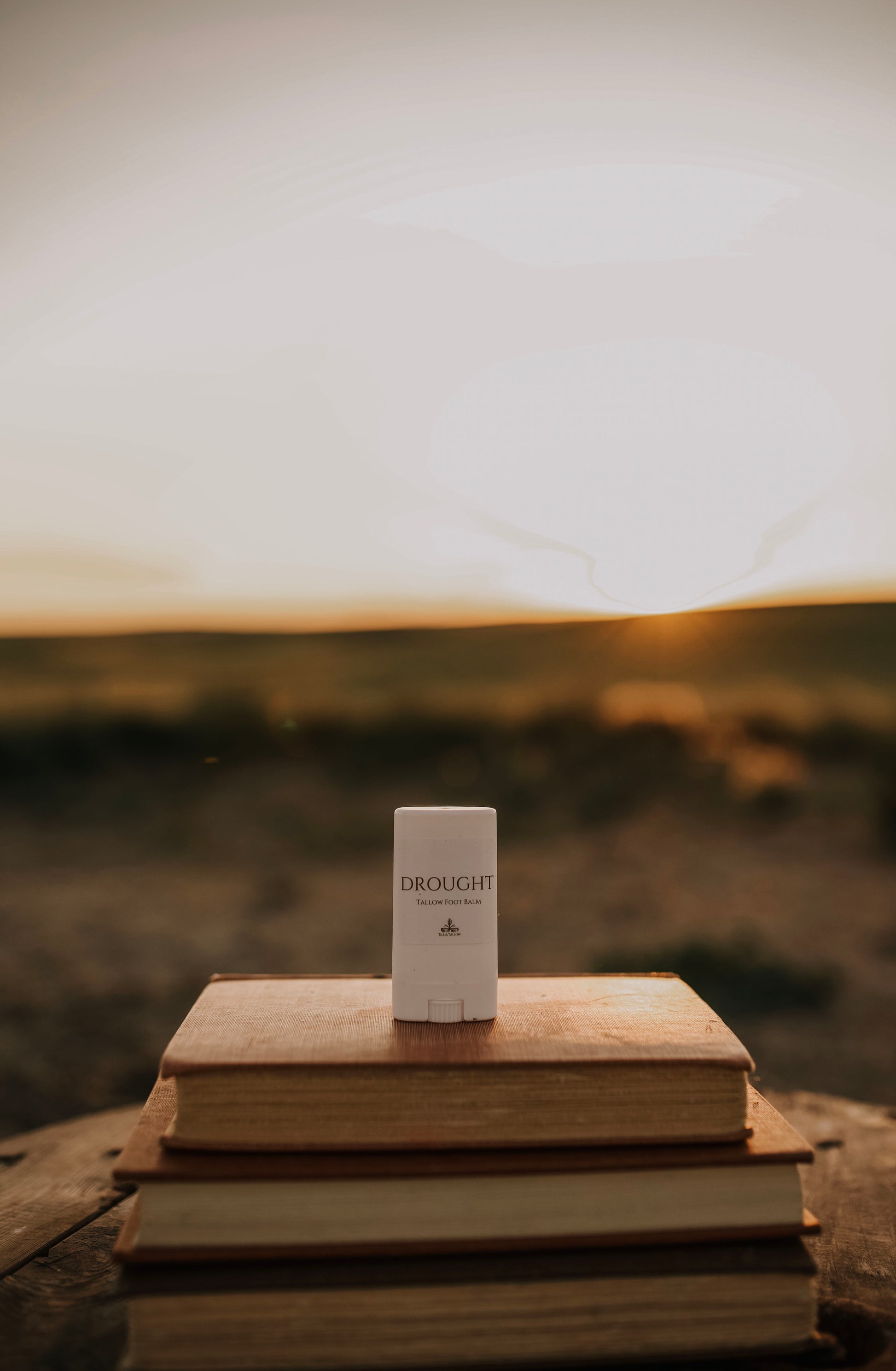 Till & Tallow Balm Drought Cracked Heel Balm in white cylindrical tube displayed on stacked vintage books outdoors with a  prairie background, handcrafted in Alberta