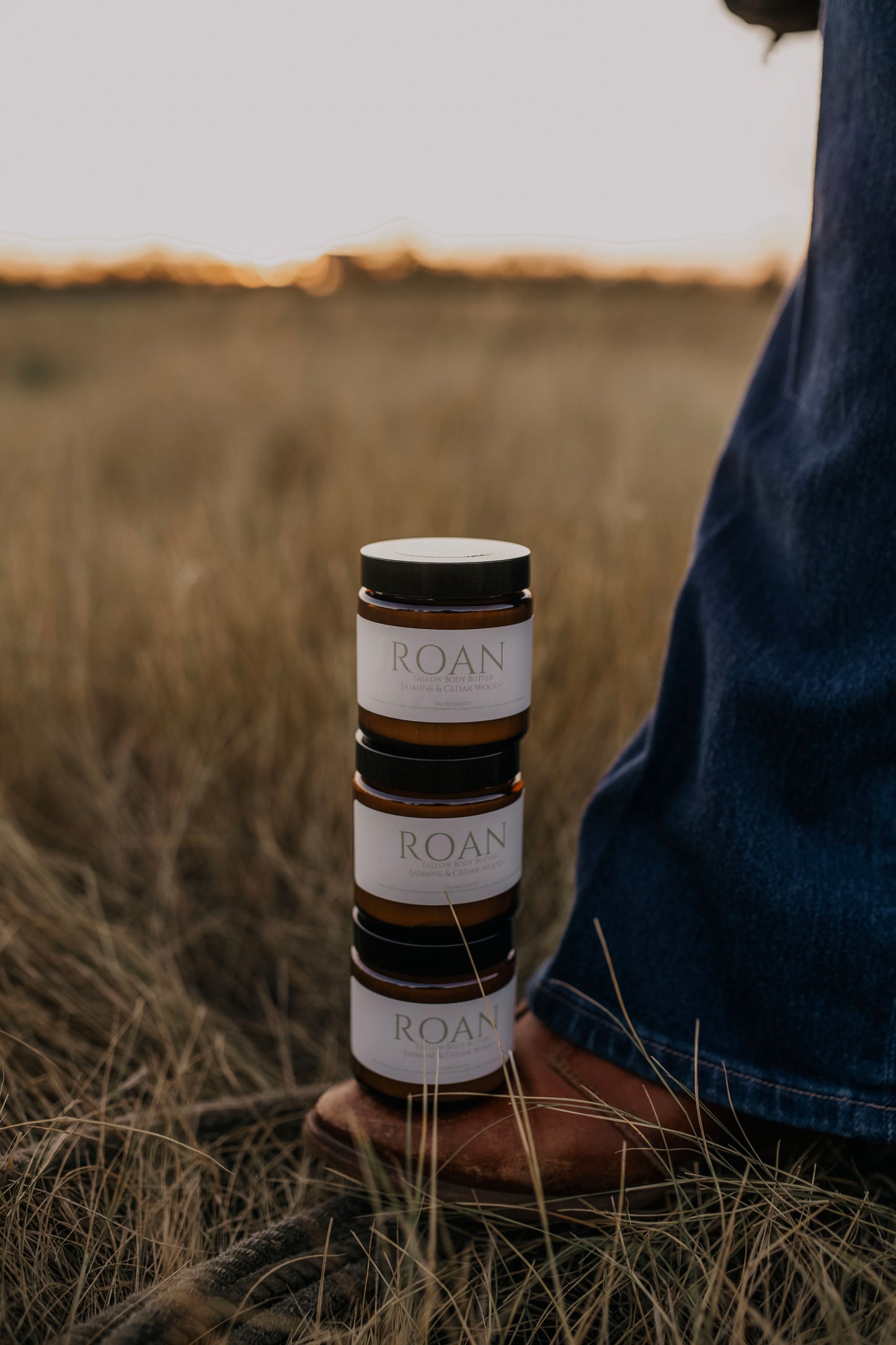 Till & Tallow Roan Body Butter jars stacked on cowboy boot in Alberta grassland field, photographed in natural light.