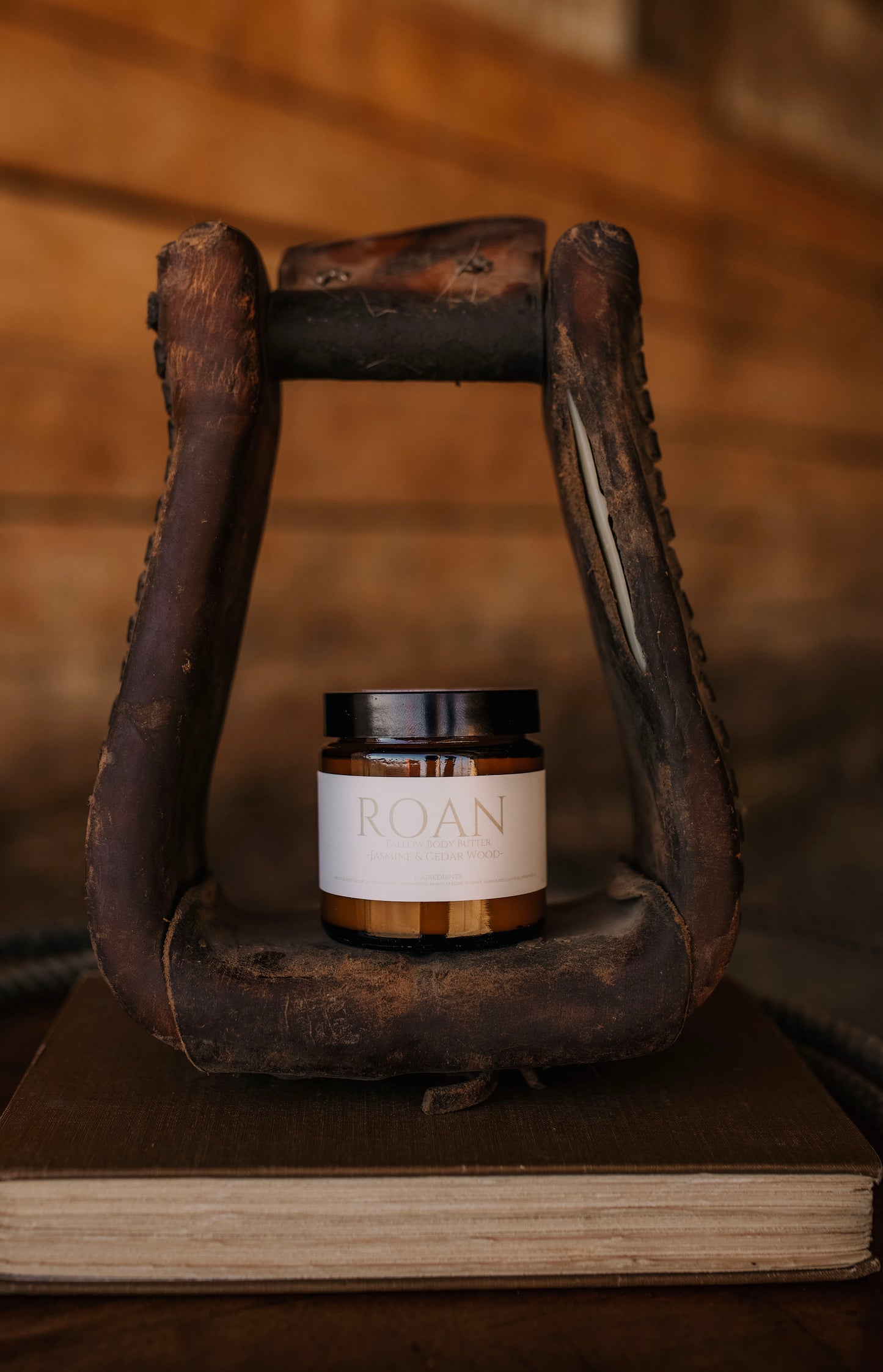 Till & Tallow Roan Body Butter in amber jar displayed inside a rustic horse stirrup on a wooden book, handcrafted in Alberta