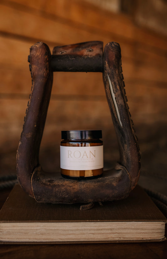 Till & Tallow Roan Body Butter in amber jar displayed inside a rustic horse stirrup on a wooden book, handcrafted in Alberta