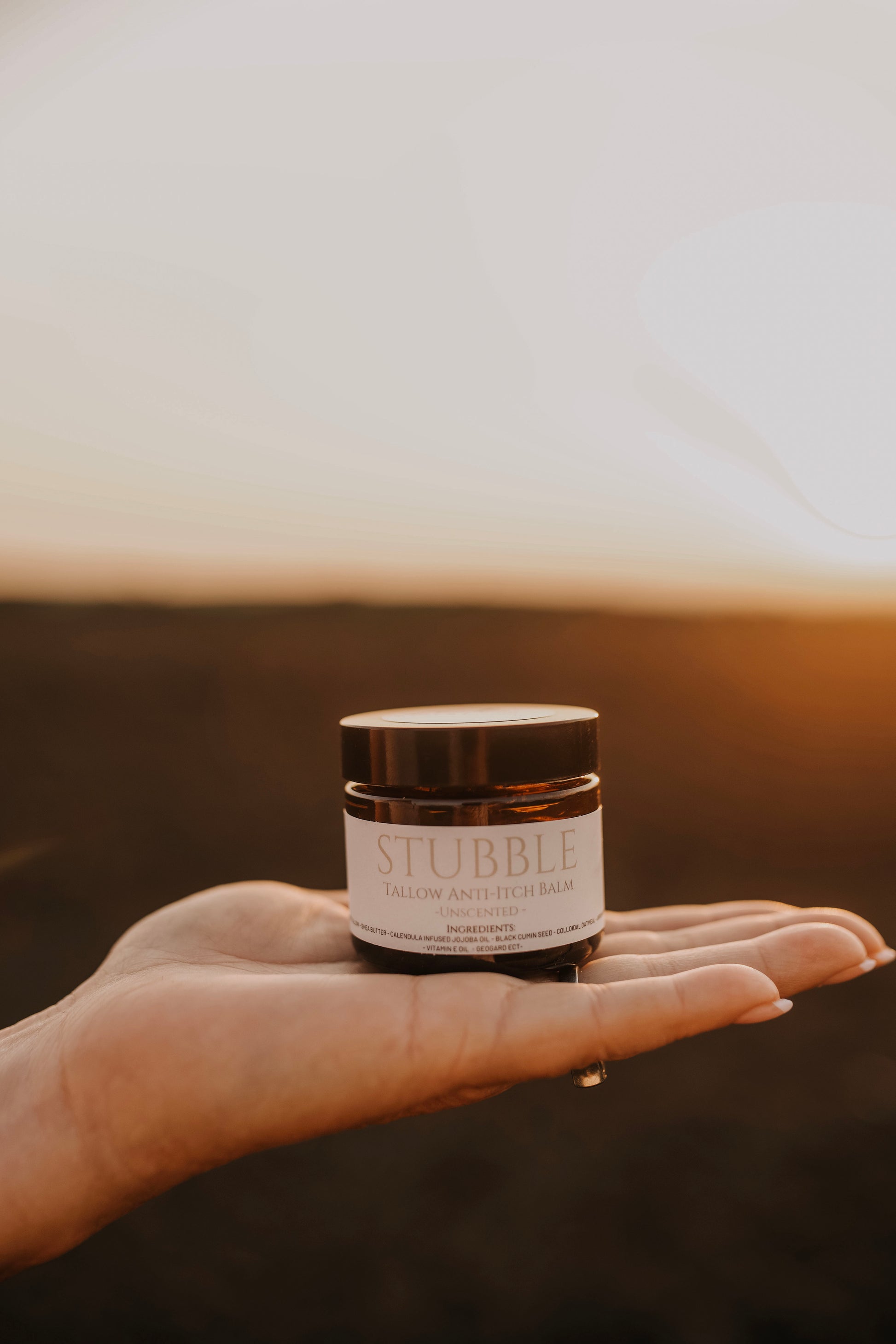 Till & Tallow Stubble Anti Itch Balm held in a open hand against a warm outdoor prairie background, handcrafted in Alberta.