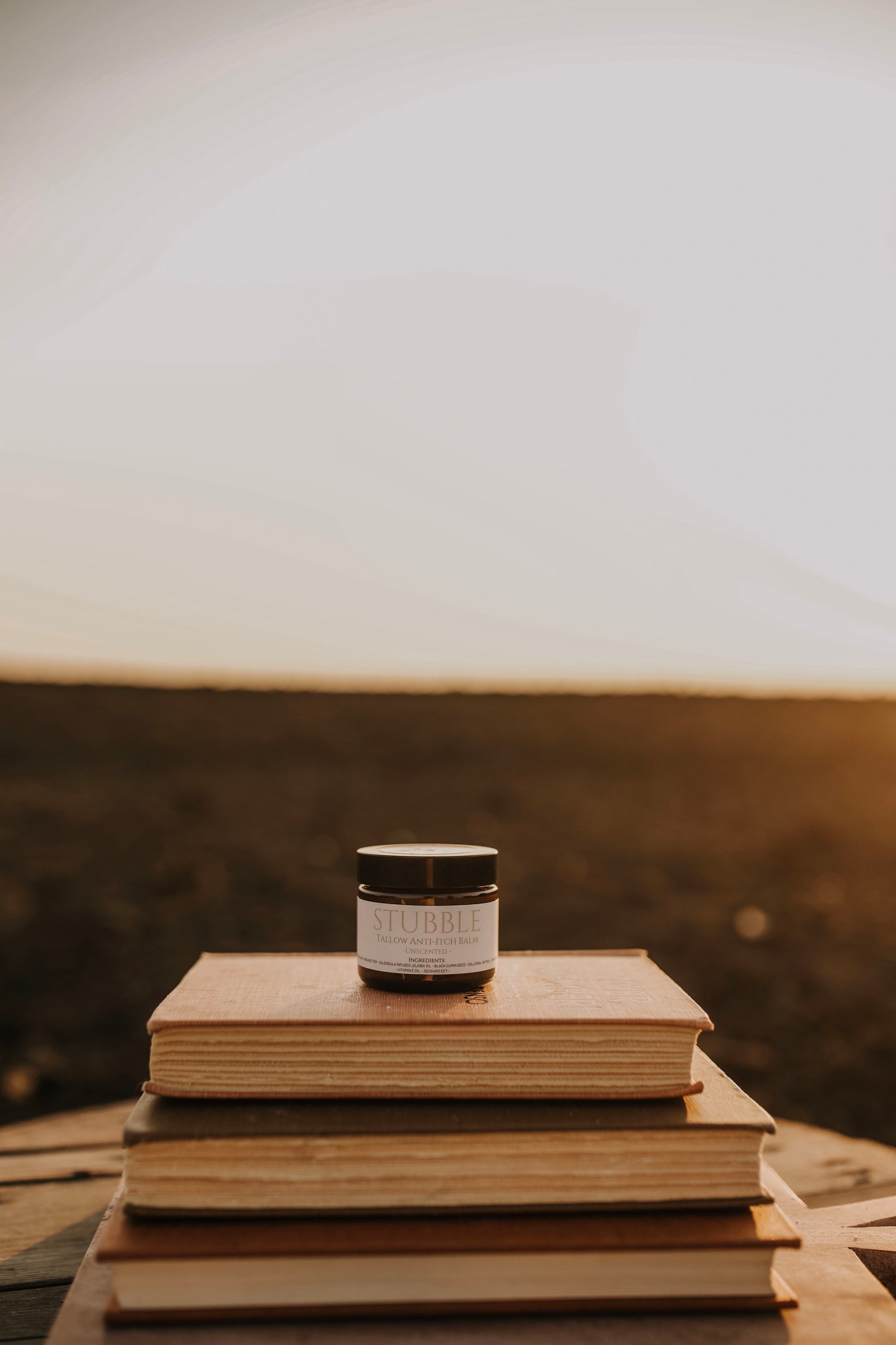 Till & Tallow Anti Itch Balm jar resting on stacked books with a warm prairie landscape in the background, handcrafted in Alberta
