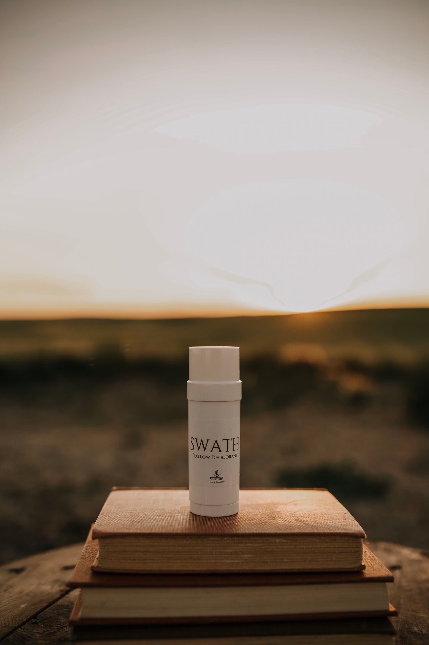 Till & Tallow SWATH natural deodorant in white tube displayed on stacked vintage books with a soft, natural outdoor background, handcrafted in Alberta.