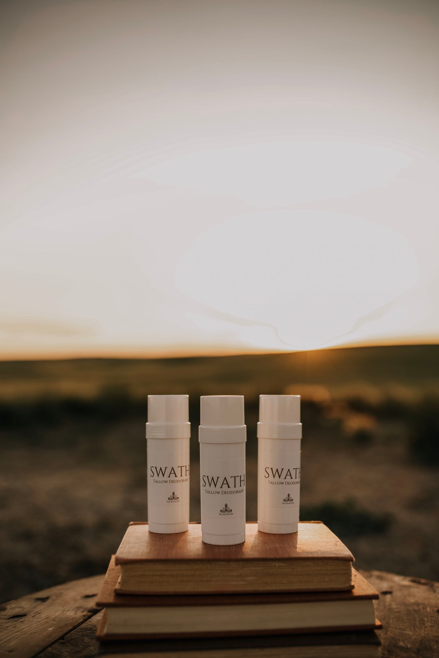 Till & Tallow SWATH natural deodorant tubes displayed on stacked vintage books outdoors with a blurred prairie background, handcrafted in Alberta.